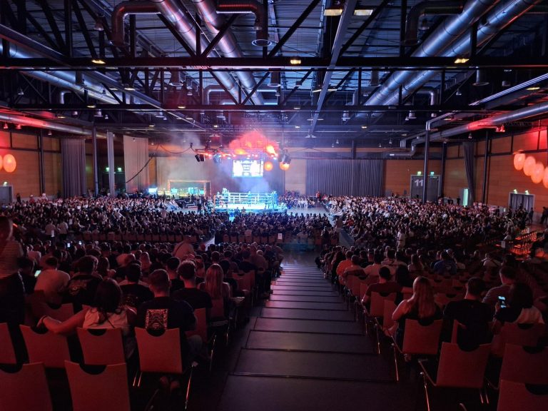 Große Menschenmenge in einer Halle während einer Veranstaltung, mit Bühnenbeleuchtung.