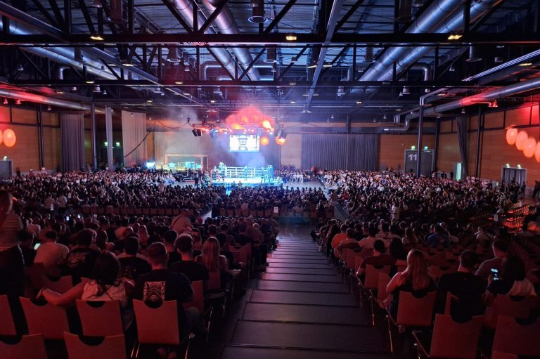 Große Menschenmenge in einer Halle während einer Veranstaltung, mit Bühnenbeleuchtung.