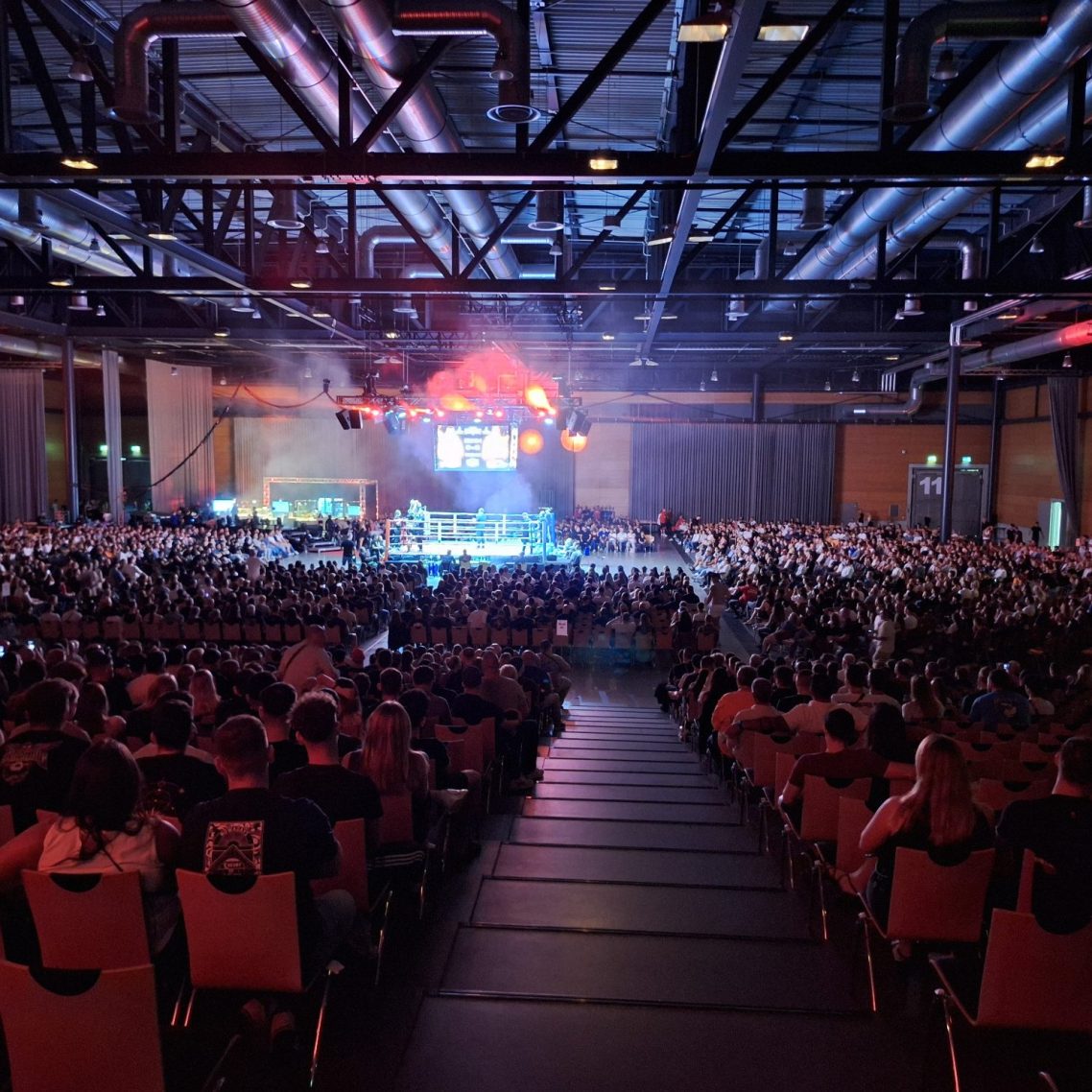 Große Menschenmenge in einer Halle während einer Veranstaltung, mit Bühnenbeleuchtung.