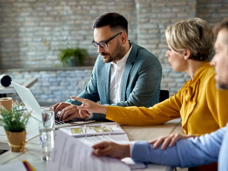Drei Personen am Tisch, diskutieren und arbeiten an einem Laptop mit Unterlagen.