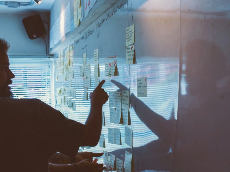 Ein Mann steht vor einem White Board mit Post-Its und zeigt auf eines dieser Post-Its.