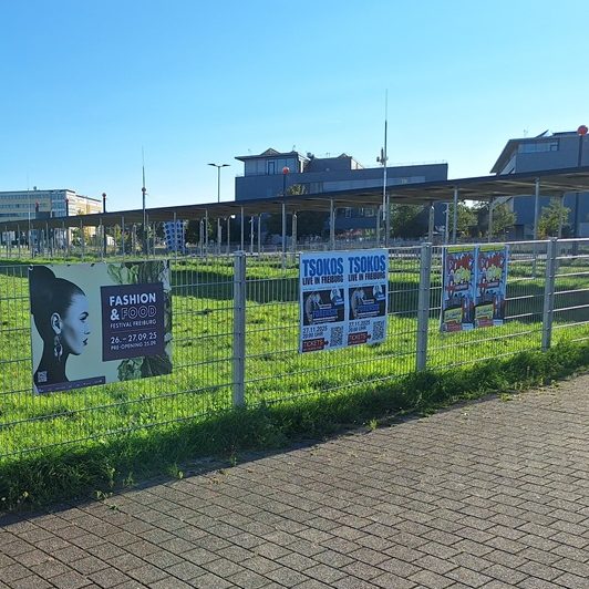 Werbetafeln an einem Zaun auf einem grasbewachsenen Grundstück unter blauem Himmel.
